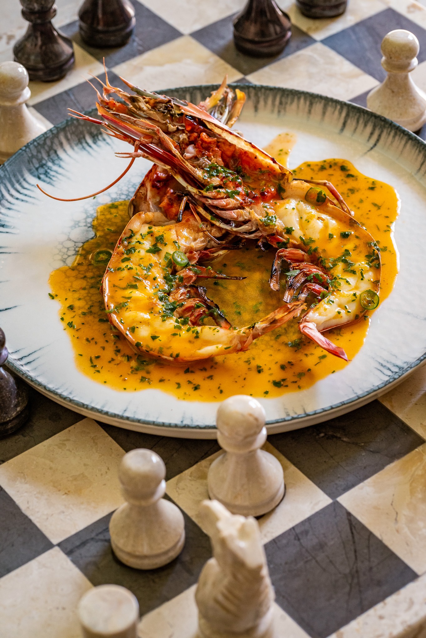 Dwa duże krewetki tygrysie, polane sosem i posypane ziołami, podane na talerzu z szarym obramowaniem, umieszczonym na szachownicy z marmurowymi figurami.