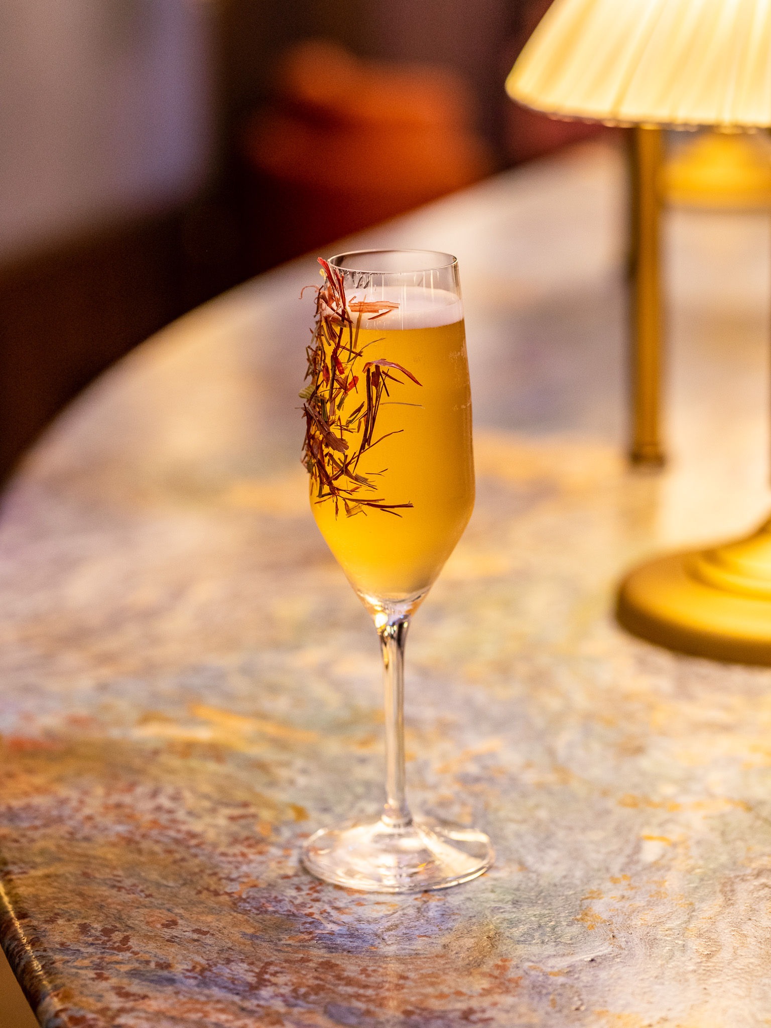 Kieliszek do szampana wypełniony złocistym napojem, ozdobiony suszonymi gałązkami, stoi na marmurowym blacie w ciepłym świetle lampki.
