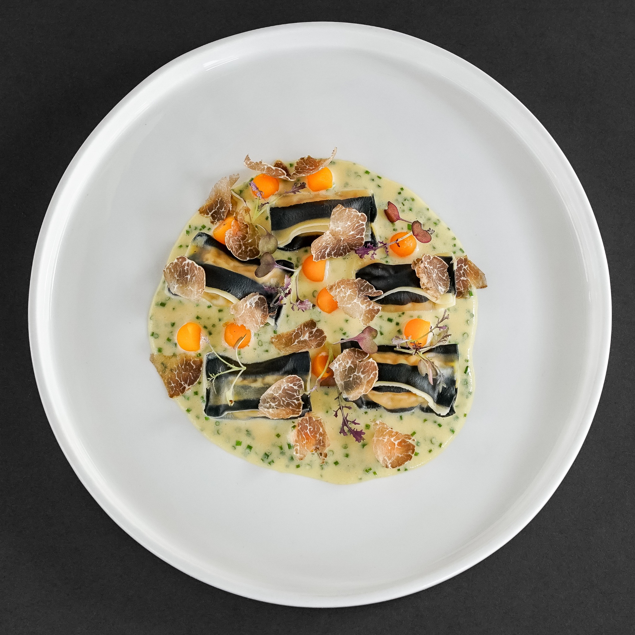 Czarno-białe ravioli z truflami i pomarańczowymi kulkami w kremowym sosie z ziołami na białym talerzu, czarne tło.