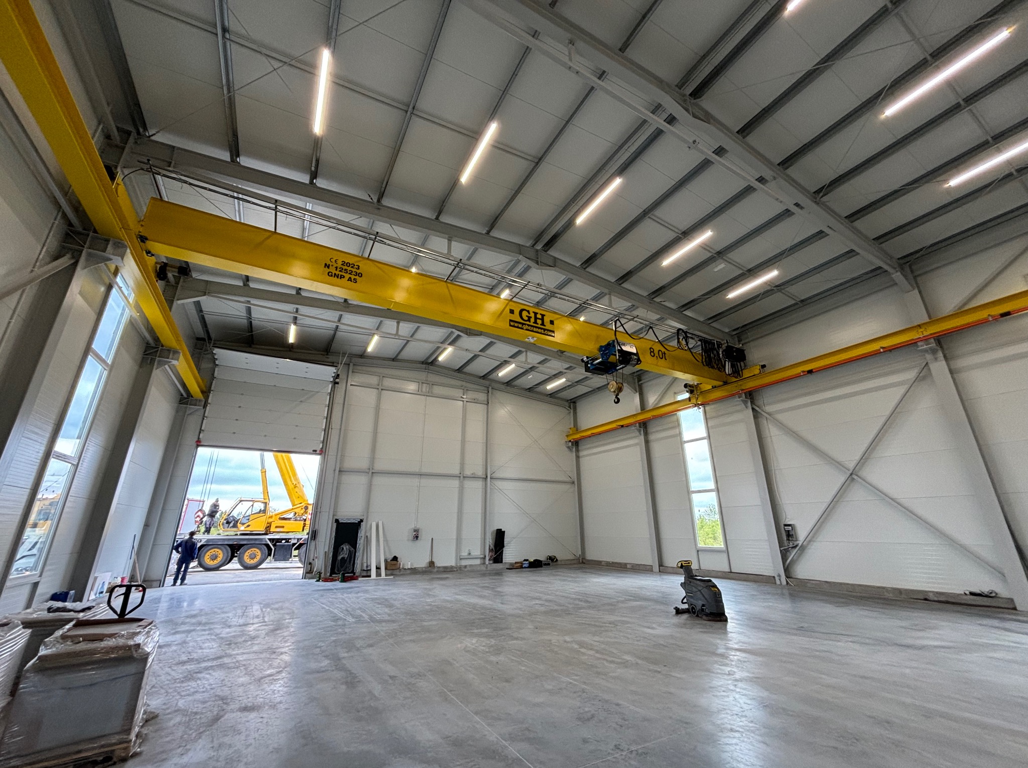 Wnętrze nowo wybudowanej hali z suwnicą, widok na otwarte wrota i pracujący dźwig na zewnątrz.