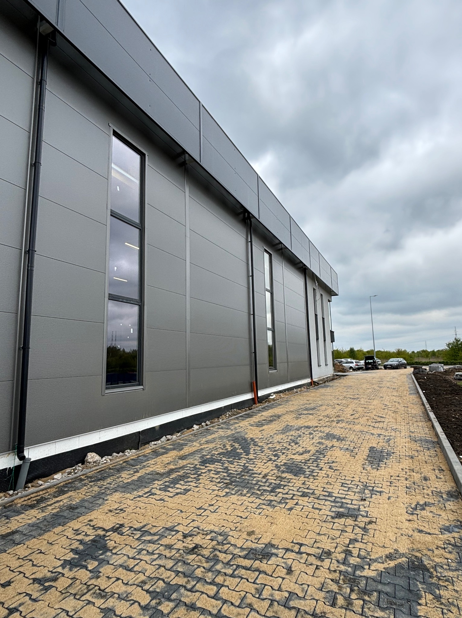 Nowoczesna hala przemysłowa z szarej płyty warstwowej, widok z zewnątrz na ułożoną kostkę brukową i pochmurne niebo.