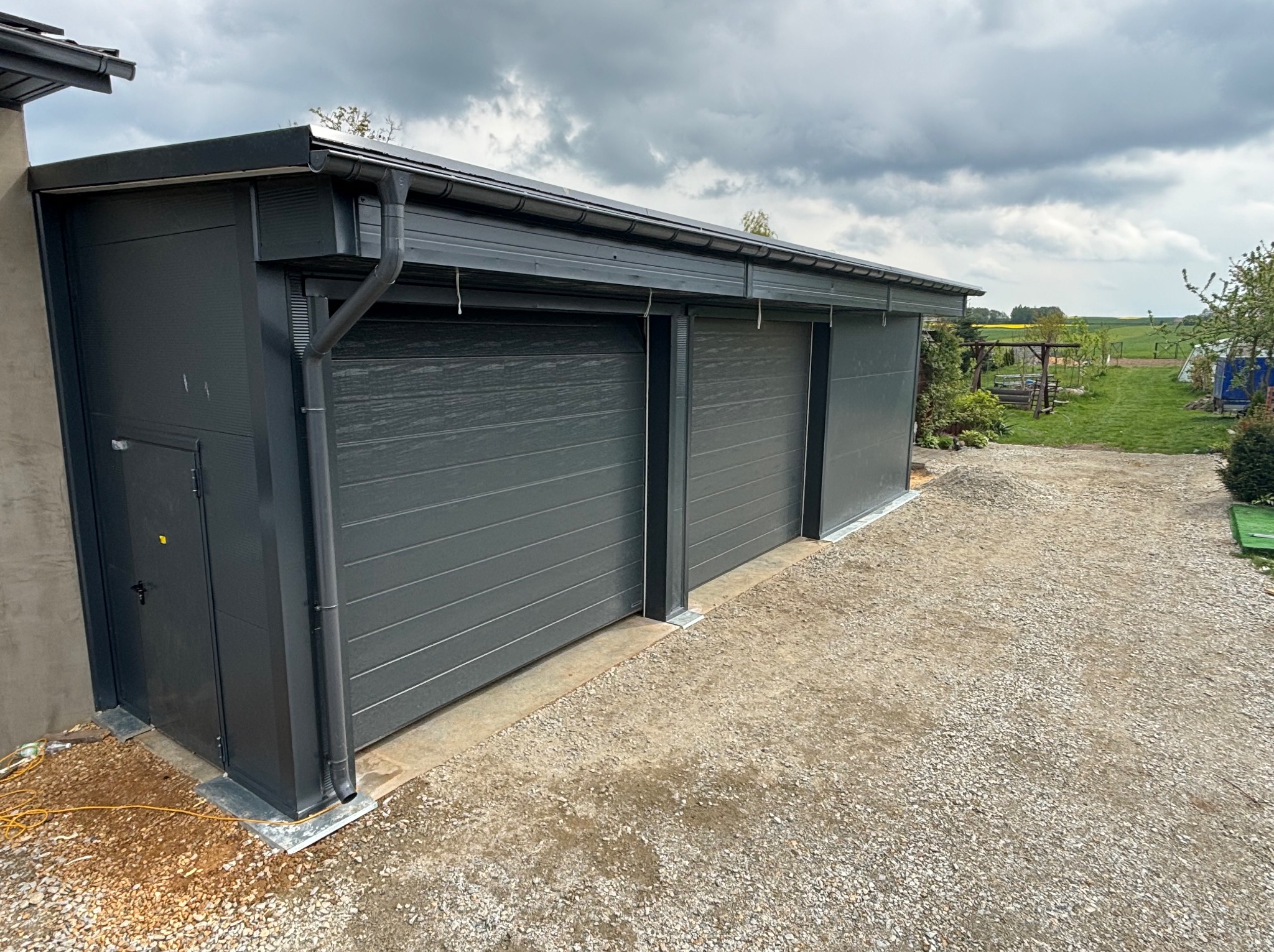 Szara hala z płyt warstwowych z dwoma bramami segmentowymi, widok z zewnątrz na żwirowy podjazd i pochmurne niebo.