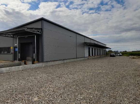 Gotowa hala przemysłowa z szarych płyt warstwowych, widoczny fragment wjazdu i rampa załadunkowa, plac wyłożony żwirem, pochmurne niebo.
