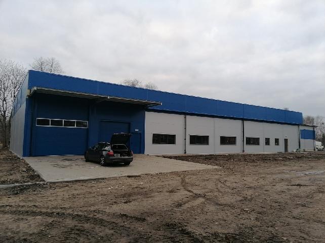 Nowoczesna hala przemysłowa z niebieskim dachem i szarymi ścianami z paneli warstwowych, zaparkowany samochód z otwartym bagażnikiem na placu budowy.