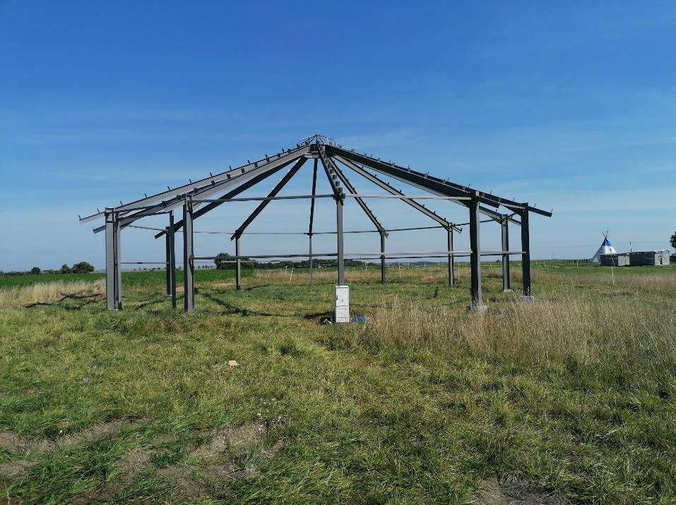 Stalowa konstrukcja hali w kształcie okręgu, w trakcie budowy na łące, widoczne elementy mocujące na dachu, w tle biały tipi.