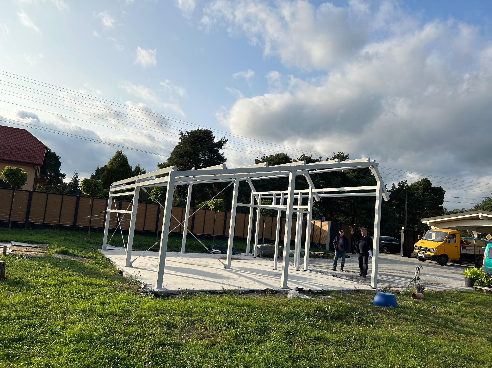 Stalowa konstrukcja hali w trakcie budowy na betonowej płycie, dwie osoby w oddali, żółty samochód dostawczy w tle, widok z trawnika.