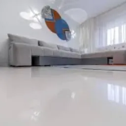 Błyszcząca, biała podłoga w salonie z narożną sofą w odcieniach szarości, abstrakcyjna dekoracja ścienna w kolorach niebieskim, pomarańczowym i szarym, oraz delikatne, białe zasłony wpadające...