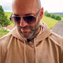 ModernTable - Mężczyzna w okularach przeciwsłonecznych i beżowej bluzie z kapturem, nagrywający wideo na tle zielonych wzgórz i domów z czerwonymi dachami.