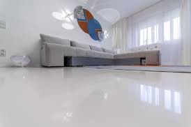 Błyszcząca, biała podłoga w salonie z narożną sofą w odcieniach szarości, abstrakcyjna dekoracja ścienna w kolorach niebieskim, pomarańczowym i szarym, oraz delikatne, białe zasłony wpadające...