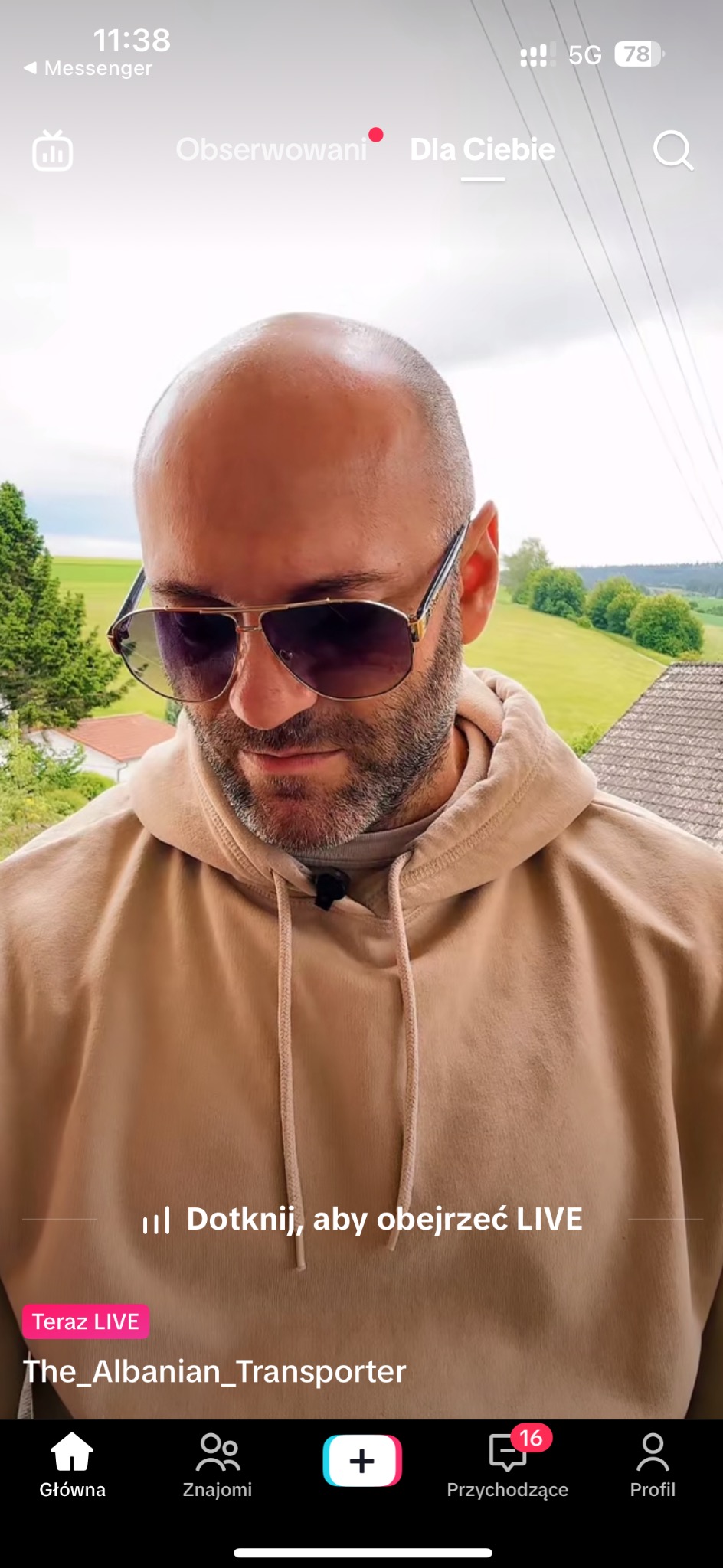 Mężczyzna w okularach przeciwsłonecznych i beżowej bluzie z kapturem, nagrywający wideo na tle zielonych wzgórz i domów z czerwonymi dachami.