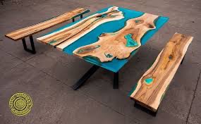 Stół i ławki z drewna i żywicy epoksydowej w kolorze turkusowym, z widocznym usłojeniem drewna i metalowymi nogami, na szarym tle. W lewym dolnym rogu logo firmy.