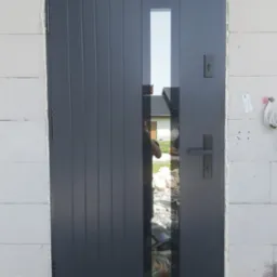 Ciemnoszare drzwi wejściowe z pionowymi żłobieniami i wąskim przeszkleniem, osadzone w murze z białych bloczków, widoczne ślady pianki montażowej wokół ościeżnicy.