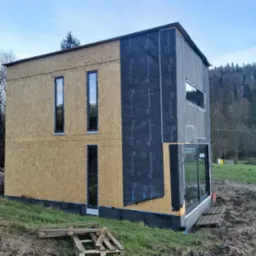 Budynek w trakcie budowy z zamontowanymi oknami PCV i membraną Eurovent na elewacji, widoczne fragmenty rusztowania i palety.