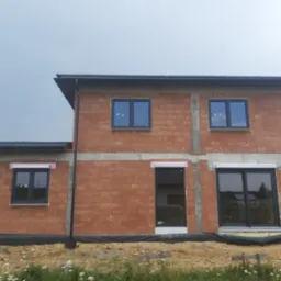 Surowy budynek z czerwonej cegły z zamontowanymi szarymi oknami i roletami zewnętrznymi w trakcie instalacji, widoczne piankowe zabezpieczenia na roletach, czarna folia izolacyjna u podstawy.