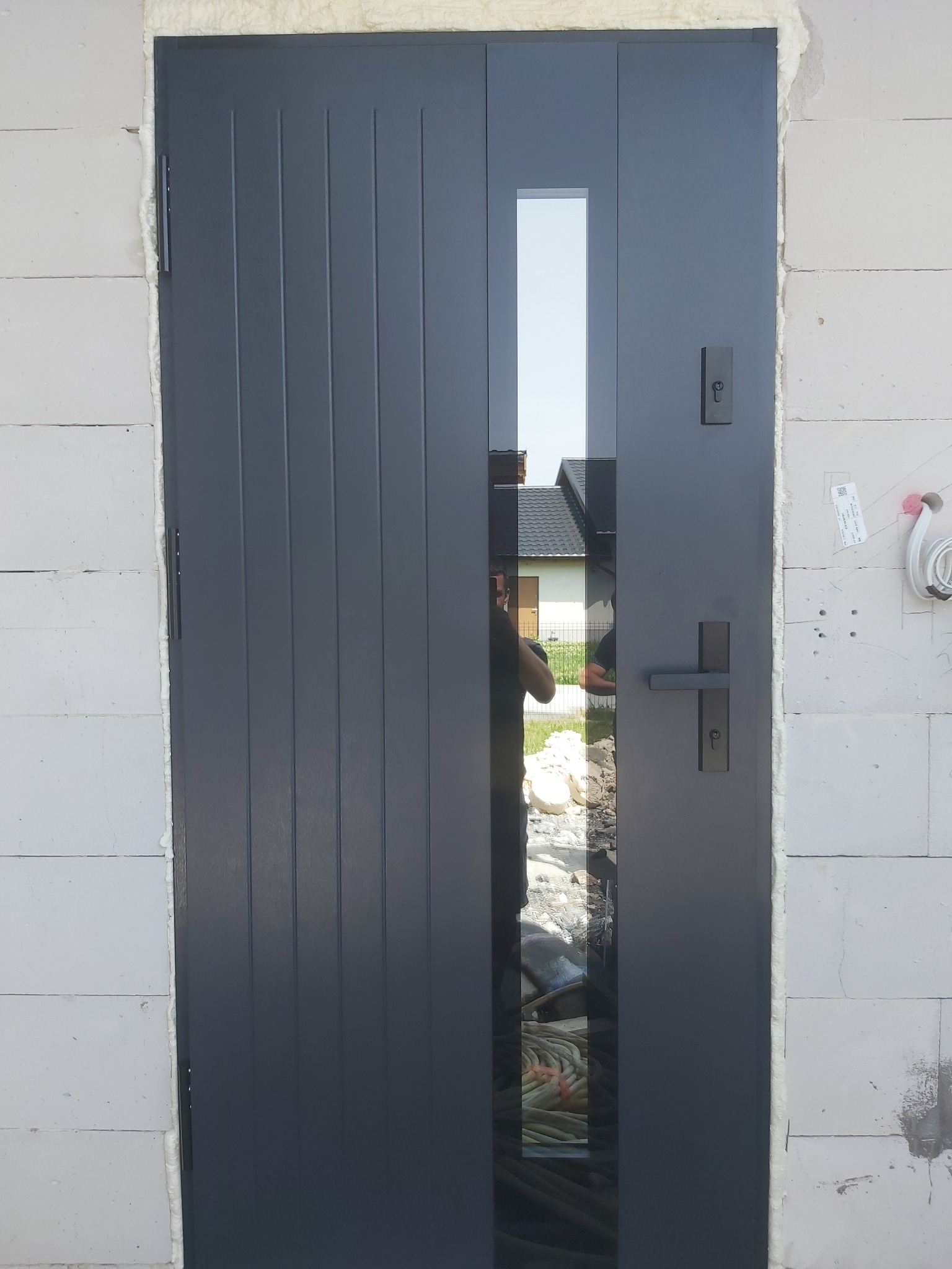 Ciemnoszare drzwi wejściowe z pionowymi żłobieniami i wąskim przeszkleniem, osadzone w murze z białych bloczków, widoczne ślady pianki montażowej wokół ościeżnicy.