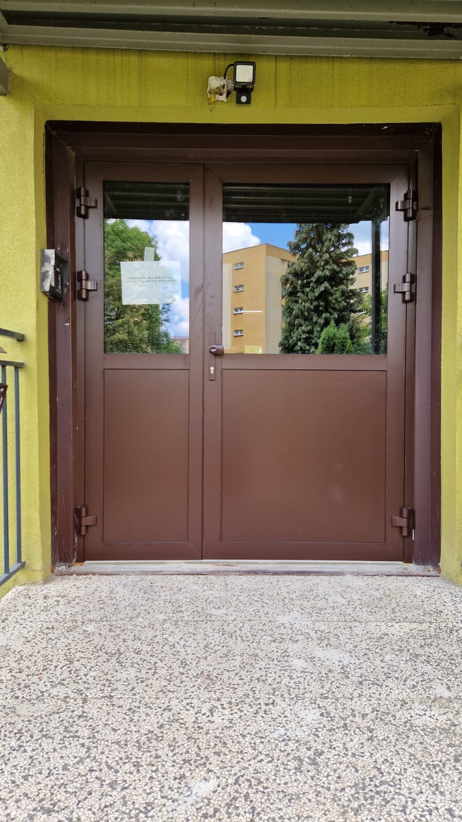 Brązowe, dwuskrzydłowe drzwi wejściowe z przeszkleniami, widok z zewnątrz na budynek i drzewo odbijające się w szybach, żółta elewacja budynku.