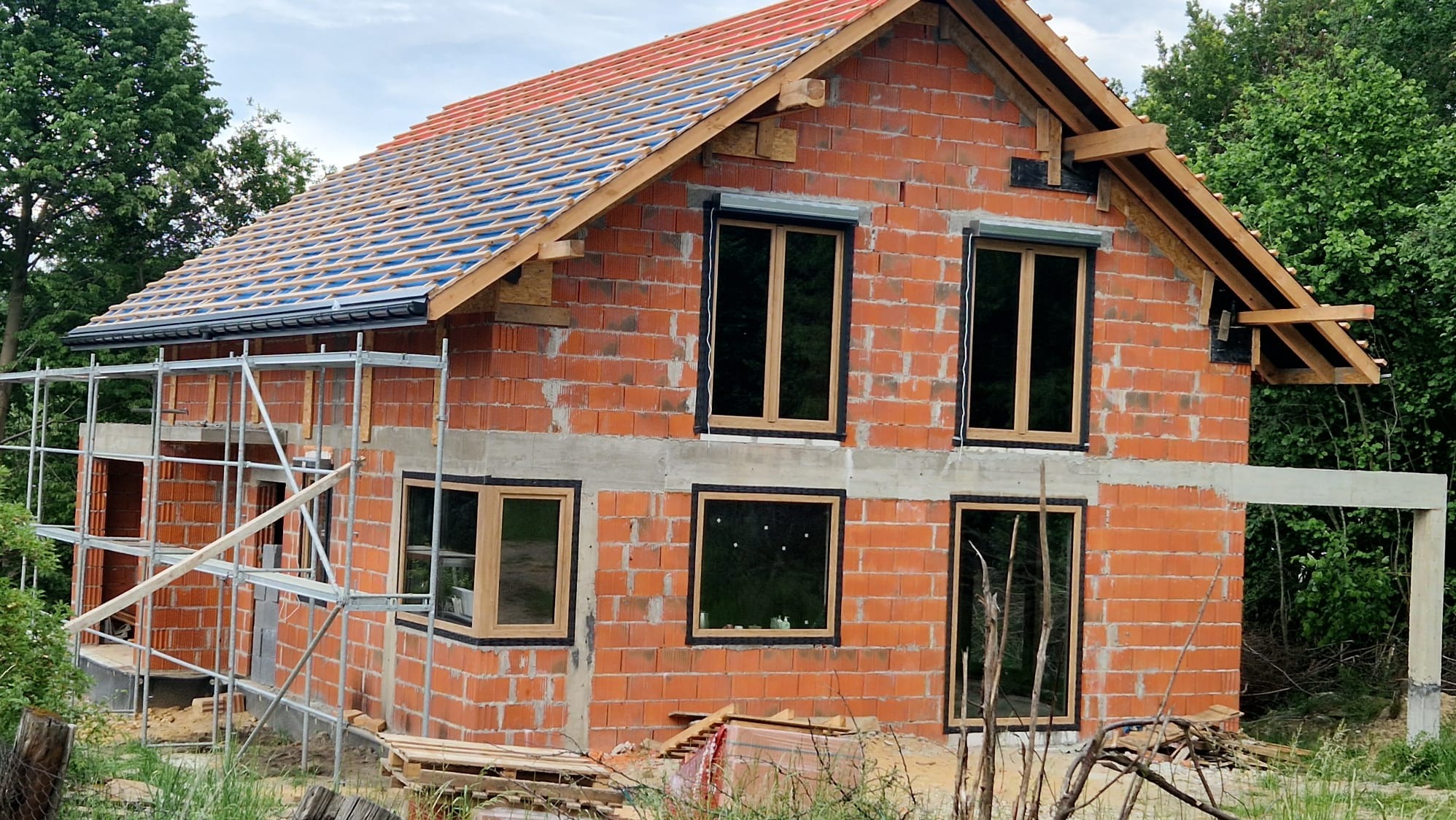 Budowa domu jednorodzinnego w stanie surowym otwartym z zamontowanymi oknami z drewnianą ramą i częściowo zamontowanymi roletami zewnętrznymi, widoczne rusztowanie i ułożona więźba dachowa.