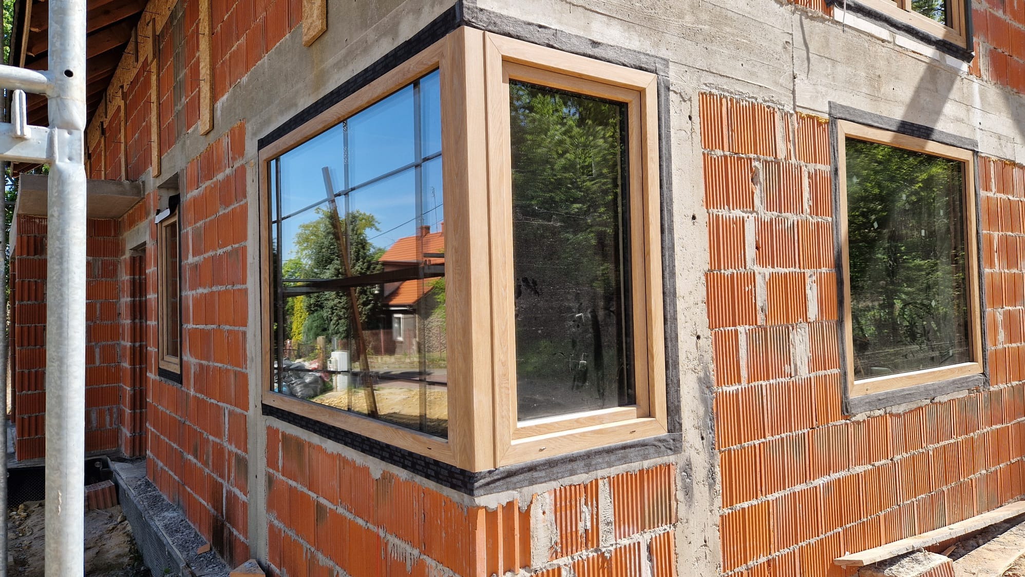 Montaż okien z drewnianą ramą w surowym budynku z cegły, widoczne detale uszczelnienia i rusztowanie w tle.