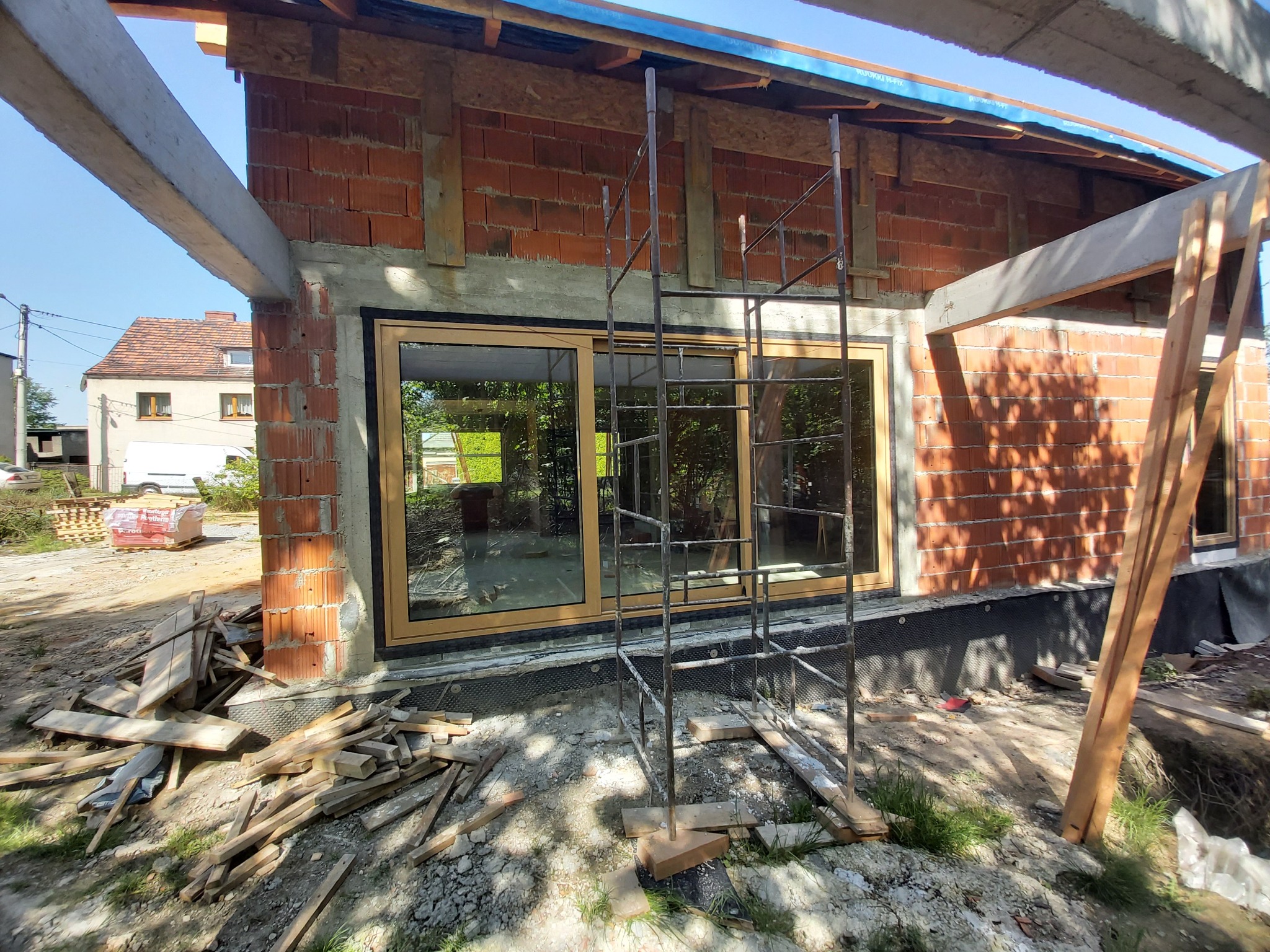 Nowe okna PCV z brązowymi ramami zamontowane w surowym murze budynku w trakcie budowy, widoczne rusztowanie i materiały budowlane.