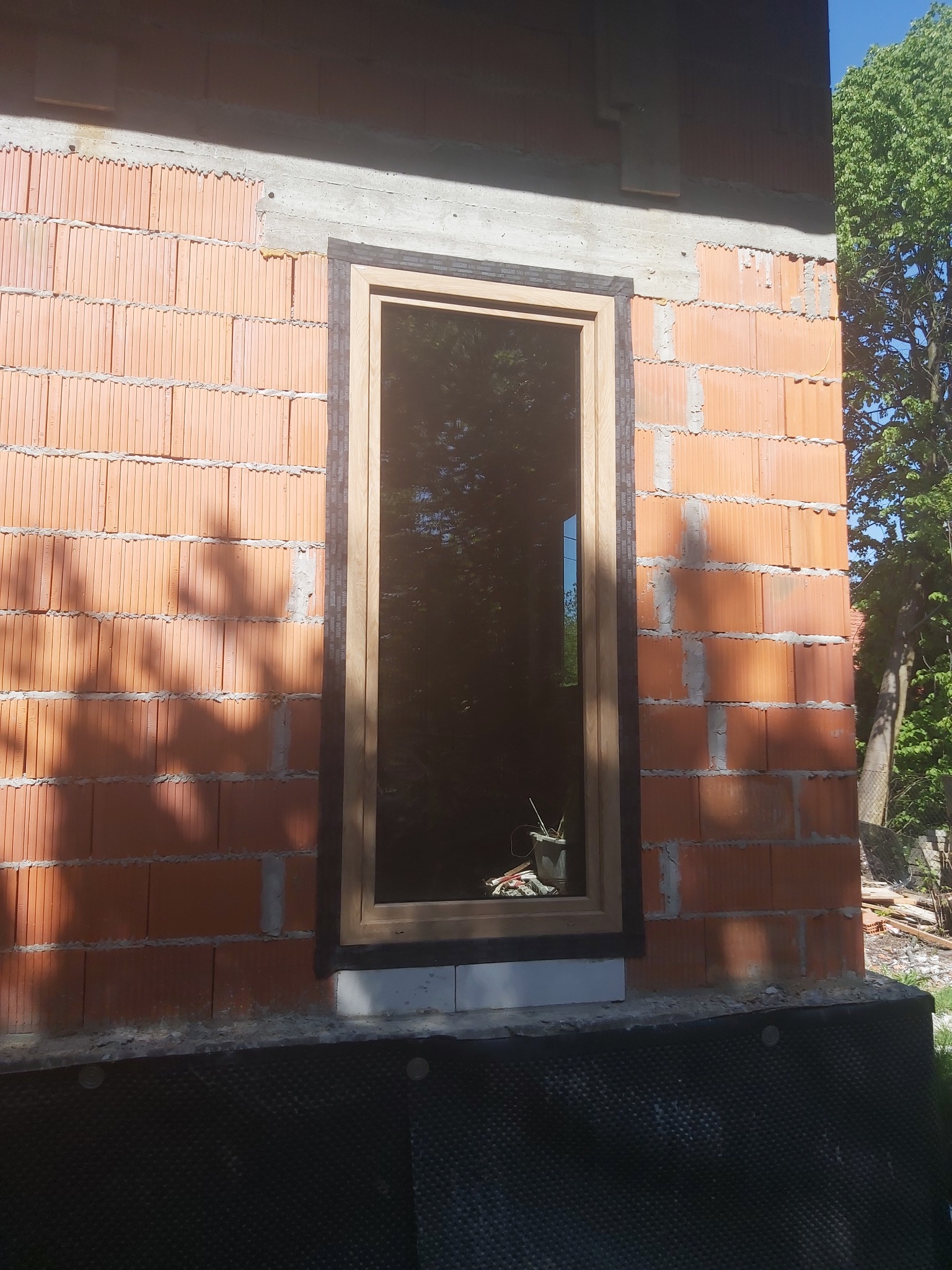 Surowy stan montażu okna z drewnianą ramą w ścianie z czerwonej cegły, widoczne elementy uszczelniające i czarna folia fundamentowa u dołu, ślady zaprawy na cegłach.