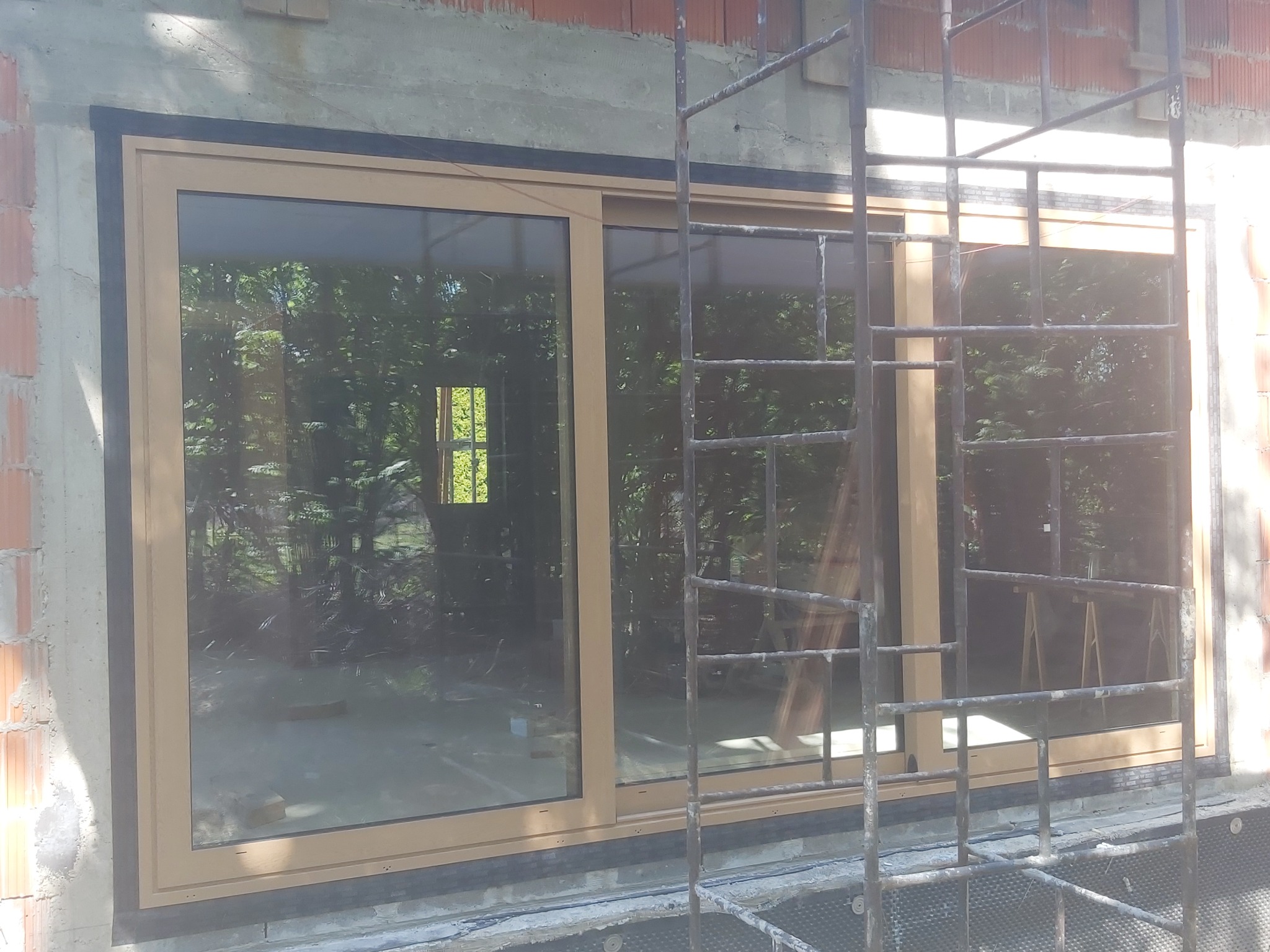 Montaż okna przesuwnego z beżową ramą w surowym murze, widoczne rusztowanie i odbicie zieleni w szybie.