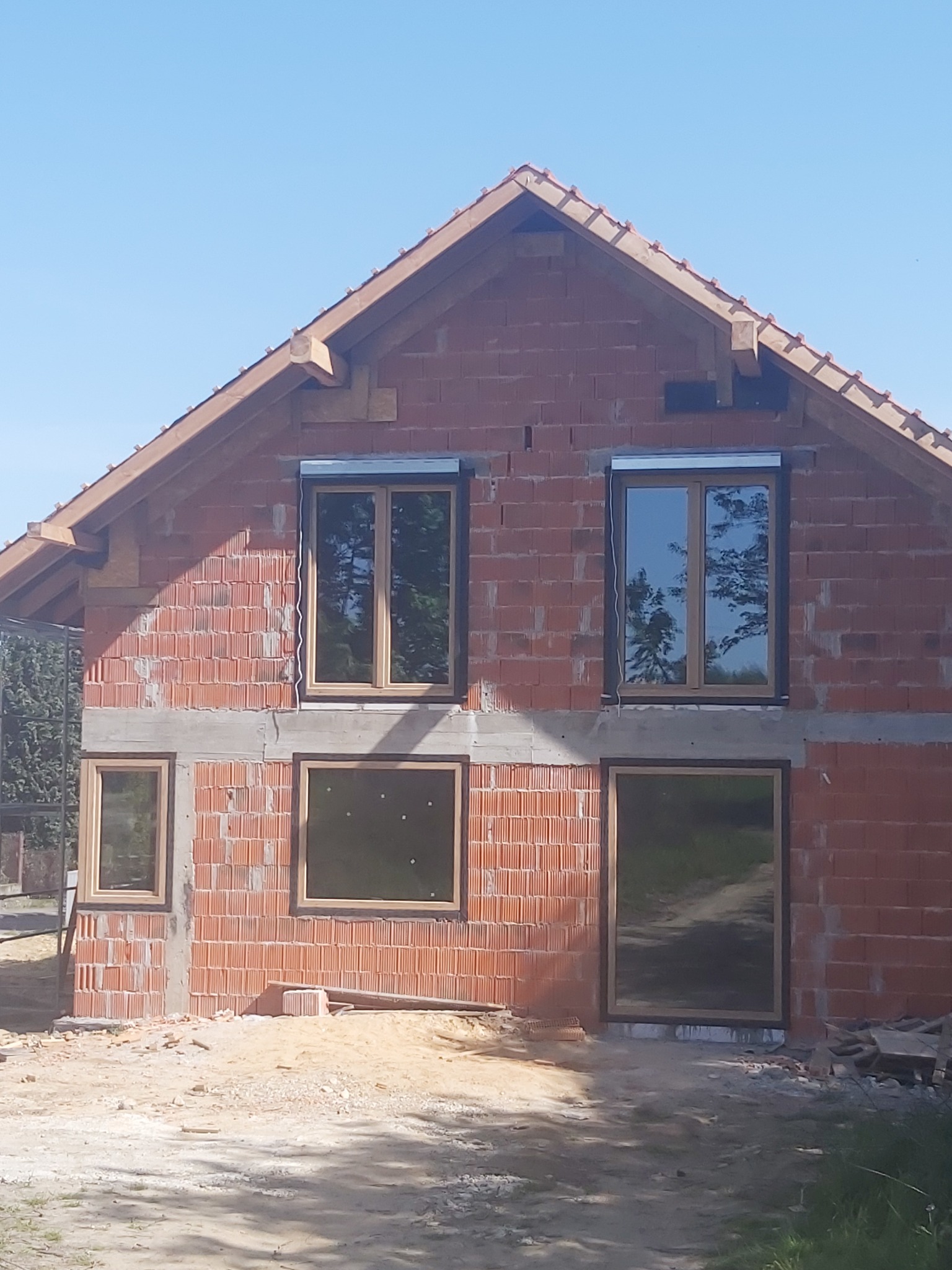 Surowy stan budynku z cegły z zamontowanymi oknami z drewnianą ramą i zewnętrznymi roletami, widoczne niedokończone prace budowlane wokół.