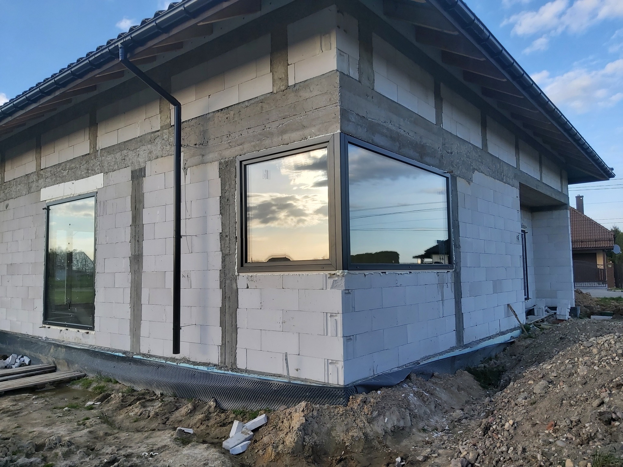 Surowy stan budynku jednorodzinnego z zamontowanymi oknami w czarnych ramach, widoczne bloczki betonowe, fundamenty, rynna spustowa i ziemia.