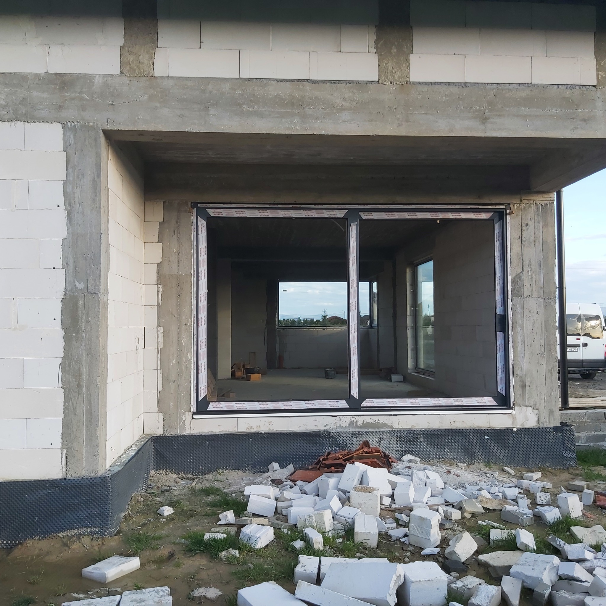 Wstawione dwuskrzydłowe okno w surowym stanie budynku z białego bloczka, widoczne gruz i fragmenty cegieł na zewnątrz.