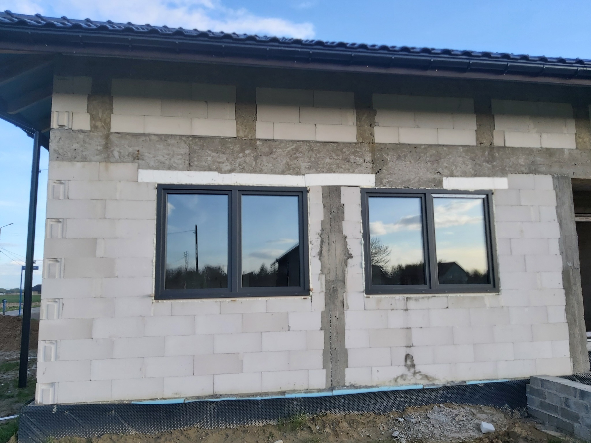 Surowy stan budynku z zamontowanymi dwoma oknami w ciemnych ramach, widoczne bloczki betonowe i fragment dachu z ciemną dachówką, odblask nieba w szybach.