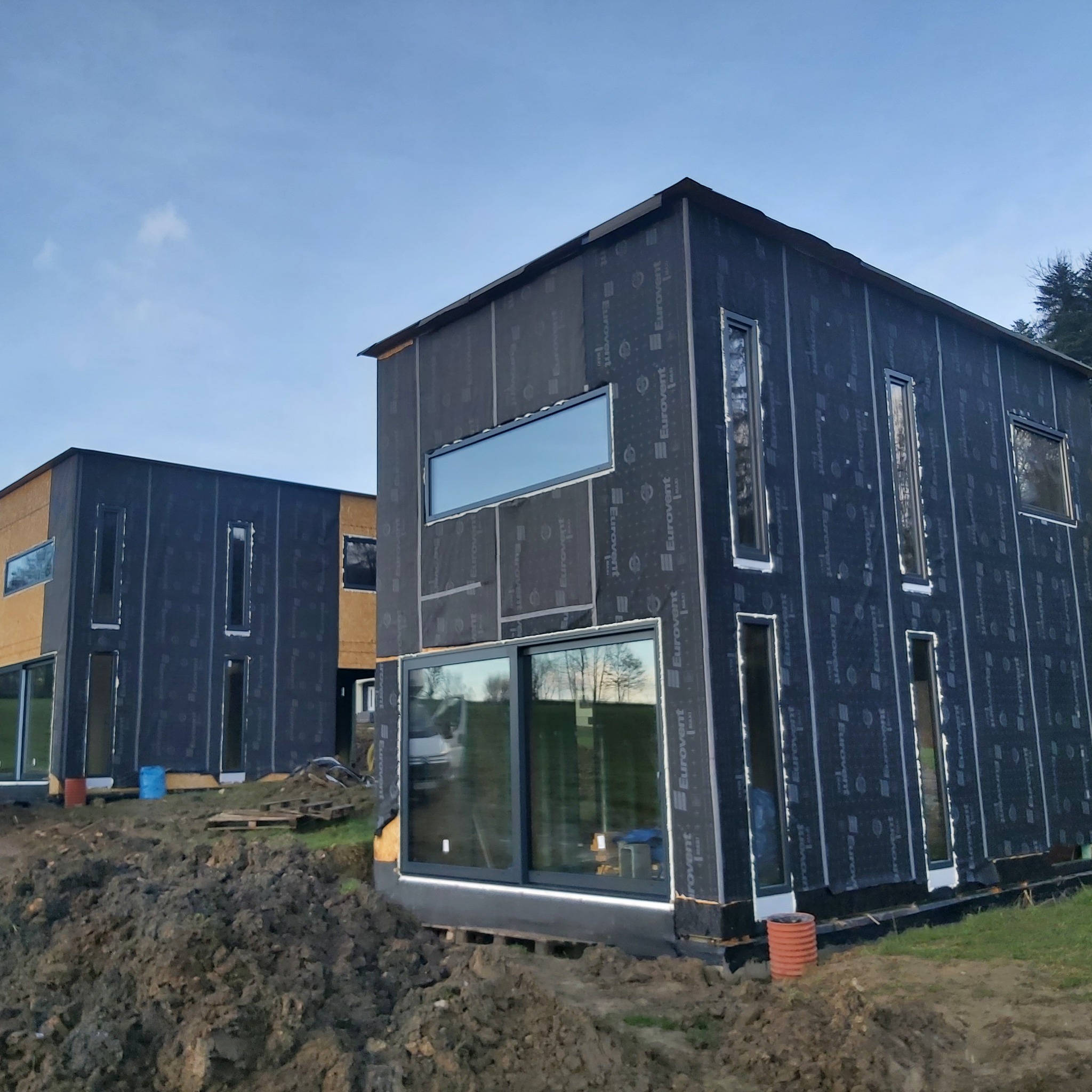 Dwa nowoczesne domy w trakcie budowy, z widocznymi oknami i membraną izolacyjną z nadrukiem, otoczone ziemią i częściowo zarośnięte trawą.