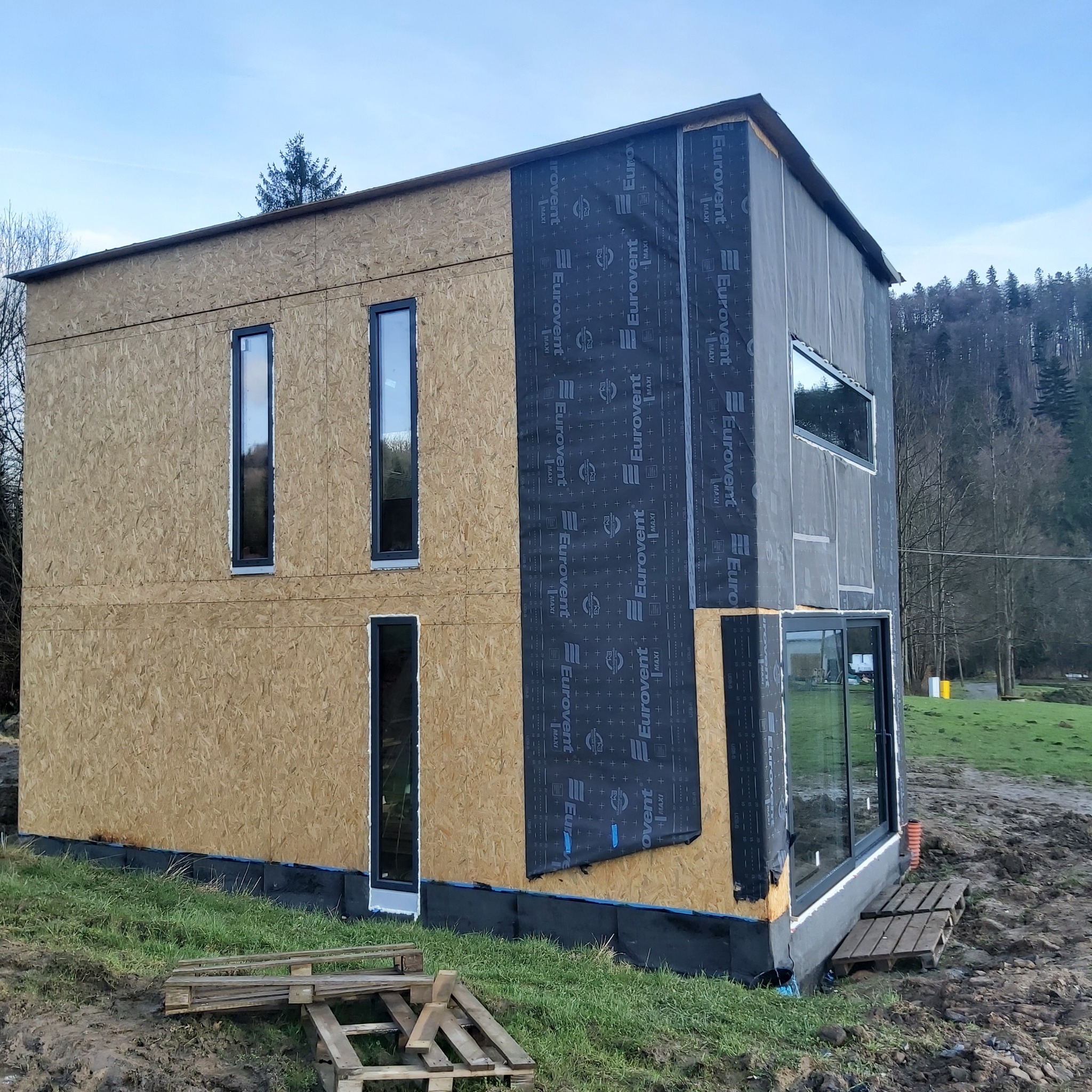 Budynek w trakcie budowy z zamontowanymi oknami PCV i membraną Eurovent na elewacji, widoczne fragmenty rusztowania i palety.