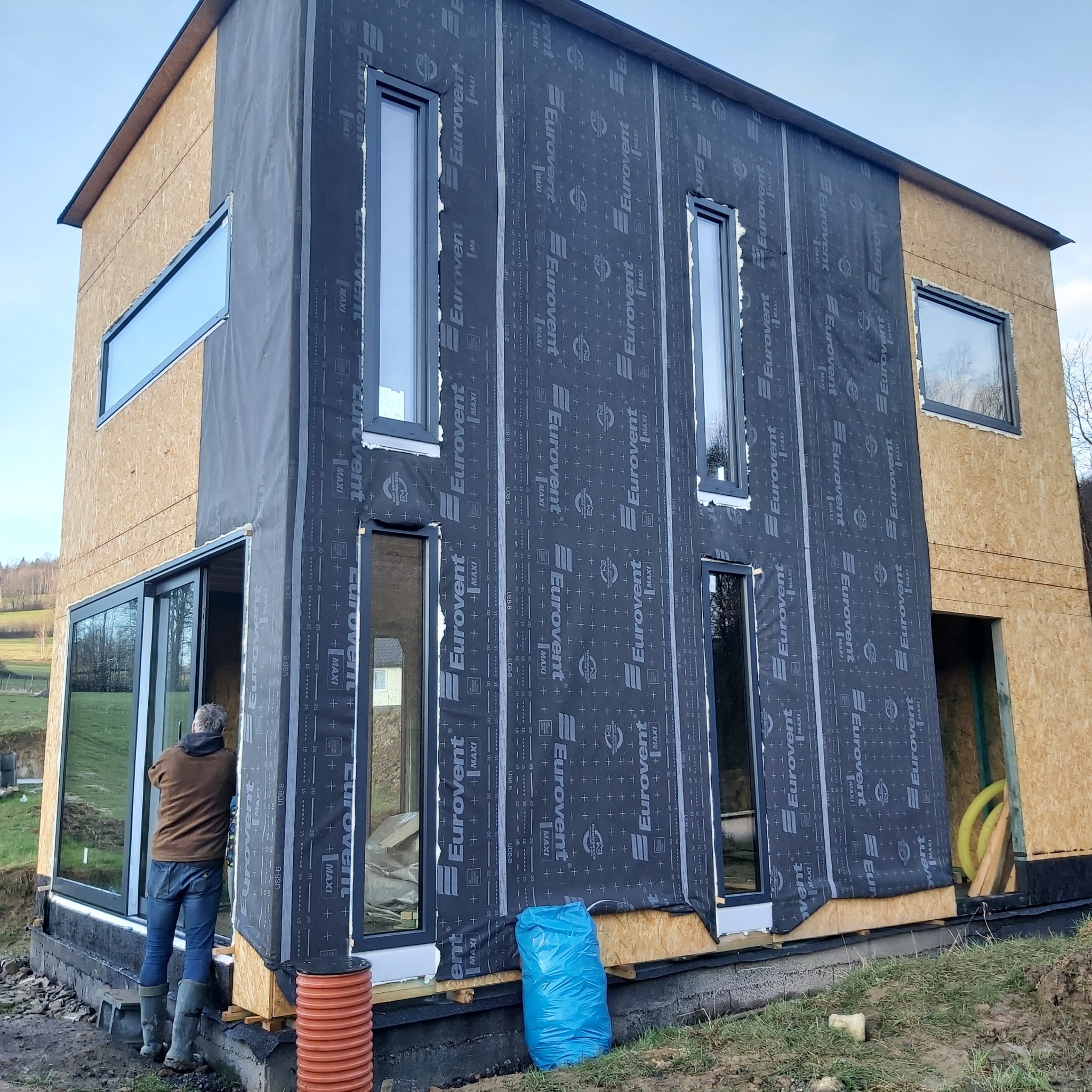 Dom w trakcie budowy, widoczne okna z czarnymi ramami zamontowane w ścianie pokrytej czarną membraną izolacyjną z nadrukiem, pracownik montuje okno przesuwne, widoczne rury wentylacyjne i niebieski...