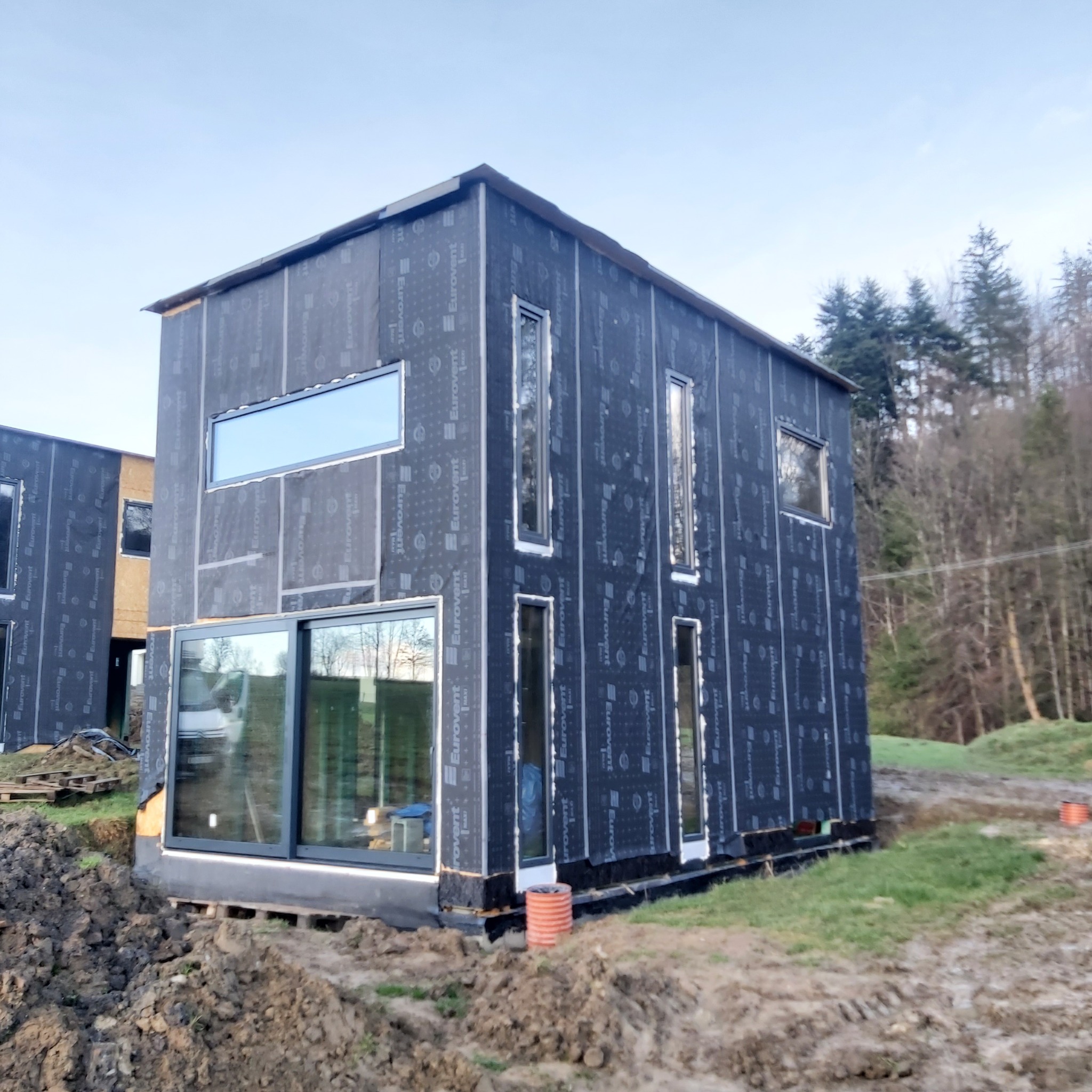 Dom w trakcie budowy, z widocznymi oknami i czarną membraną izolacyjną z nadrukiem Eurovent, na nierównym terenie z drzewami w tle.
