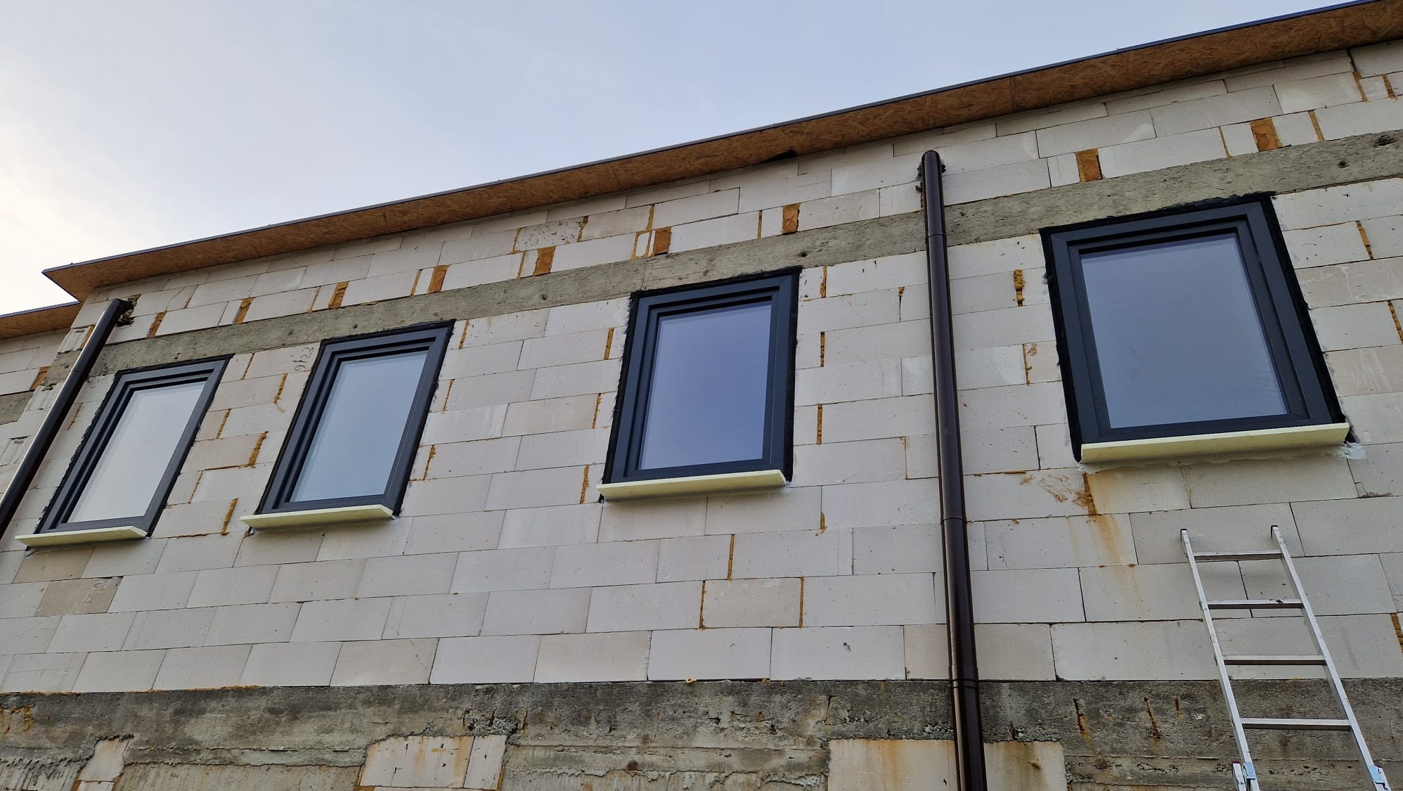 Nowo zainstalowane czarne ramy okienne w surowym murze z betonu komórkowego, widoczne elementy ocieplenia pod oknami, drabina oparta o ścianę.
