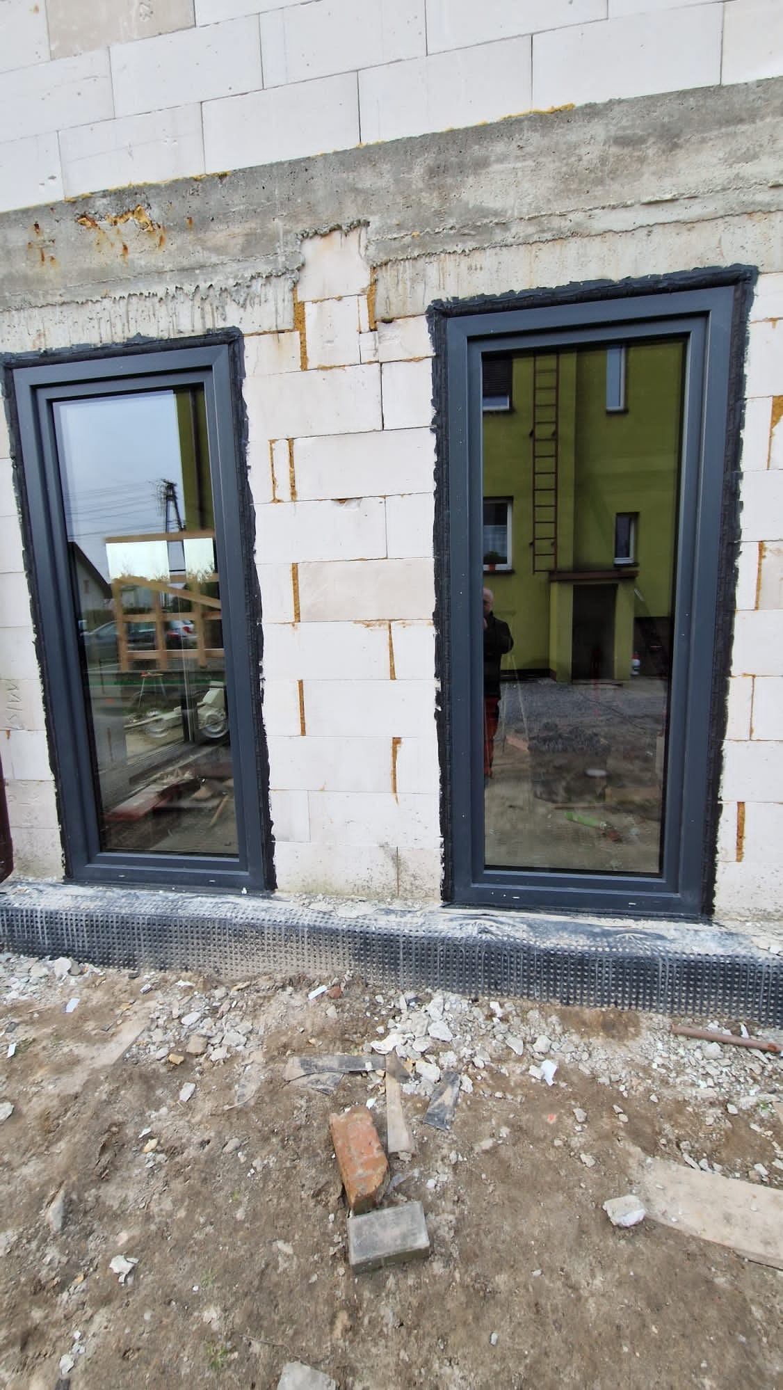 Montaż okien PCV w surowym stanie budynku w Tarnowskich Górach, widoczne dwa okna z szarymi ramami osadzone w ścianie z białych bloczków, wokół okien widoczna pianka montażowa, teren wokół budynku...