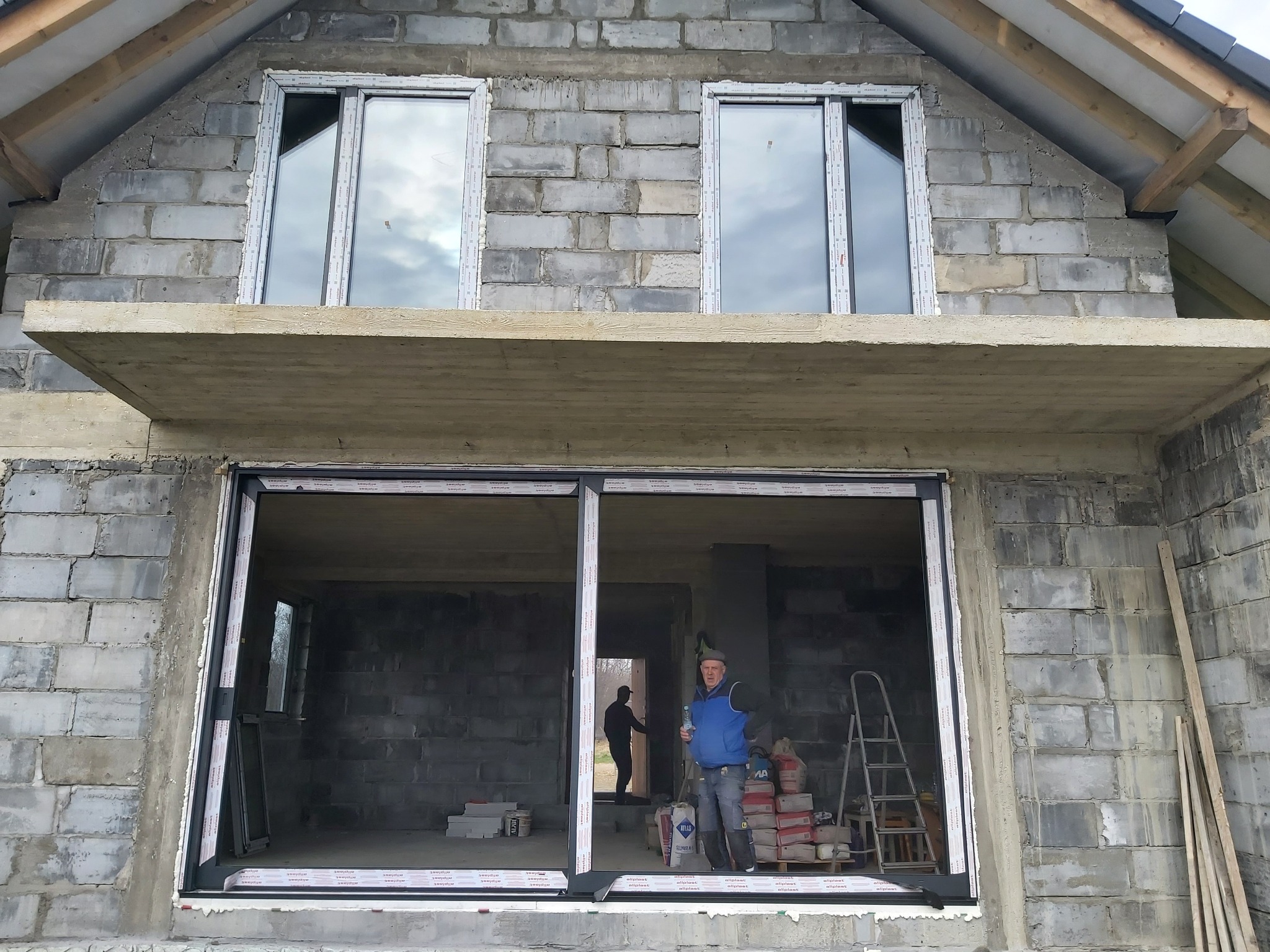 Surowy stan budynku z zamontowanymi oknami na poddaszu i dużym oknem przesuwnym na parterze. Widoczny pracownik z butelką wody i drabina oparta o ścianę. W tle drugi pracownik.