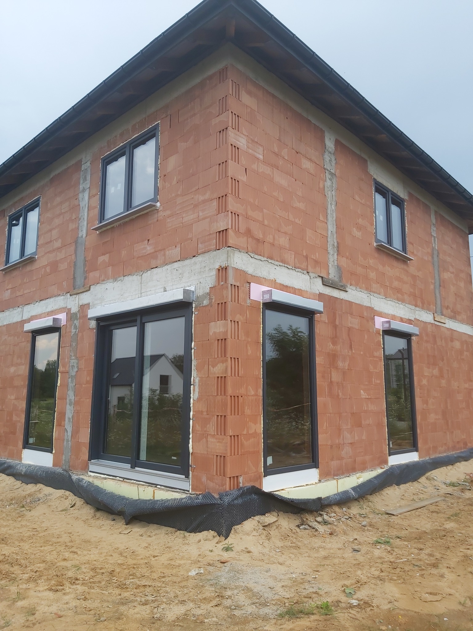 Surowy budynek z cegły z zamontowanymi oknami PCV z szarymi ramami i częściowo zamontowanymi roletami zewnętrznymi, widoczne ocieplenie i zabezpieczenie fundamentów.