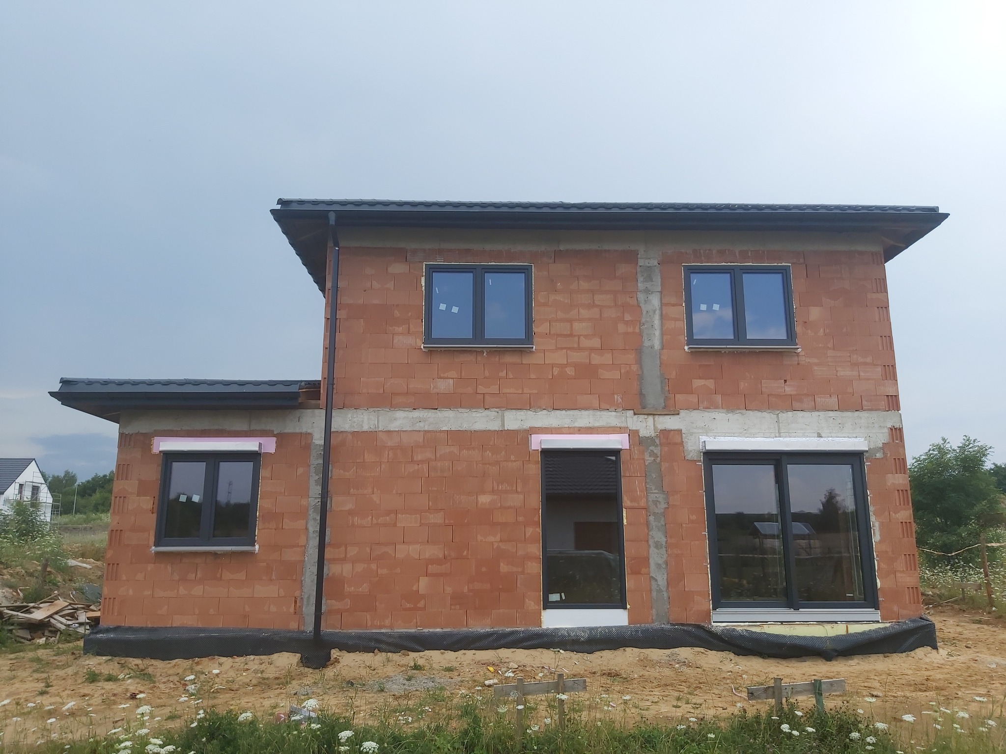 Surowy budynek z czerwonej cegły z zamontowanymi szarymi oknami i roletami zewnętrznymi w trakcie instalacji, widoczne piankowe zabezpieczenia na roletach, czarna folia izolacyjna u podstawy.