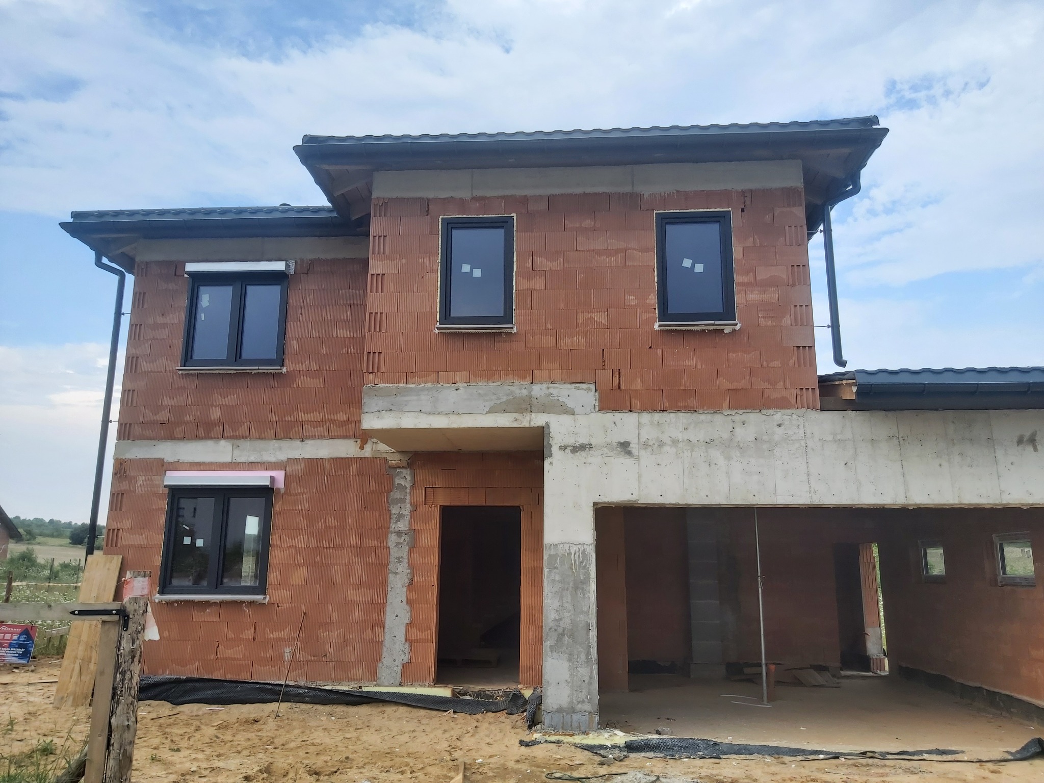 Surowy budynek w trakcie budowy z zamontowanymi oknami w czarnych ramach i roletami zewnętrznymi, widoczne pustaki i betonowe nadproża, teren wokół budynku piaszczysty.