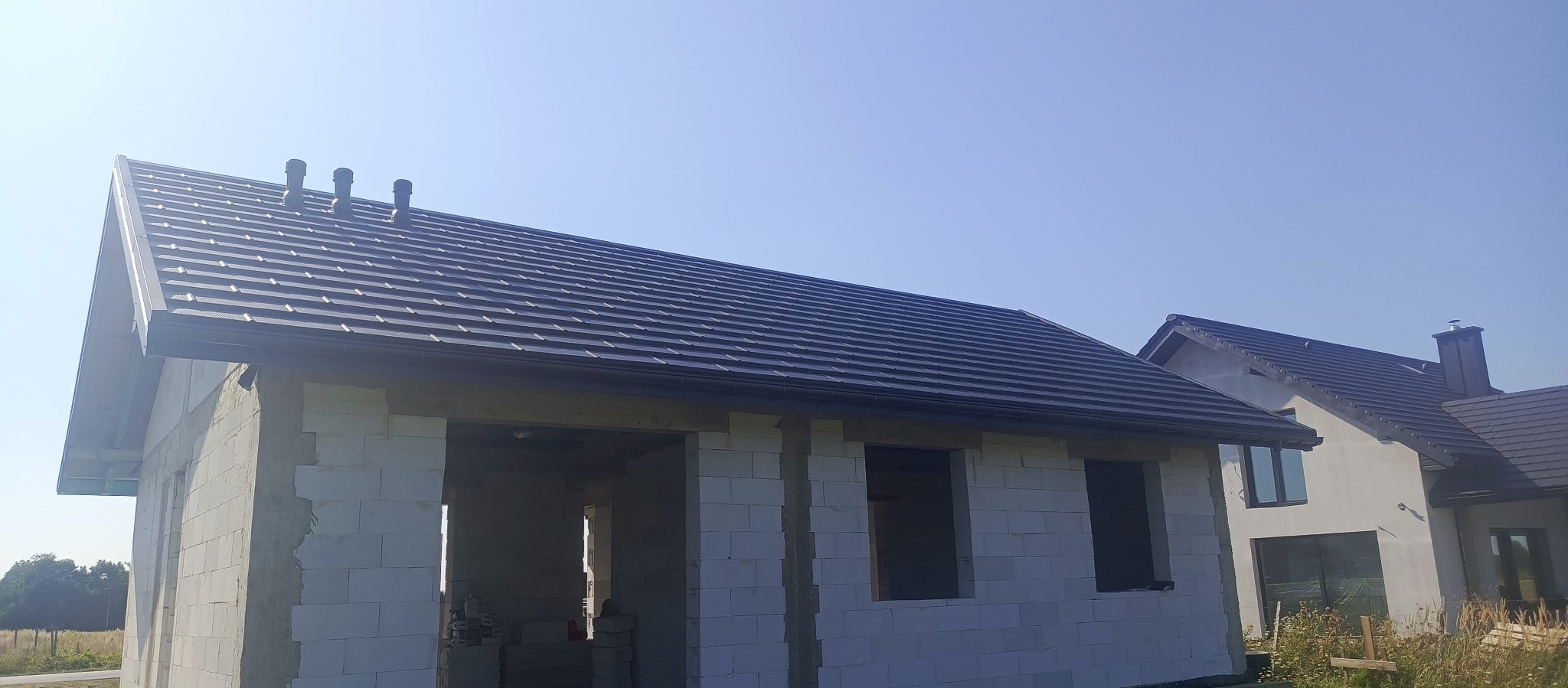 Surowy dom z białego pustaka z ciemnym, nowym dachem z dachówki ceramicznej. Widoczne okna bez szyb. W tle drugi dom z podobnym dachem i kominem.