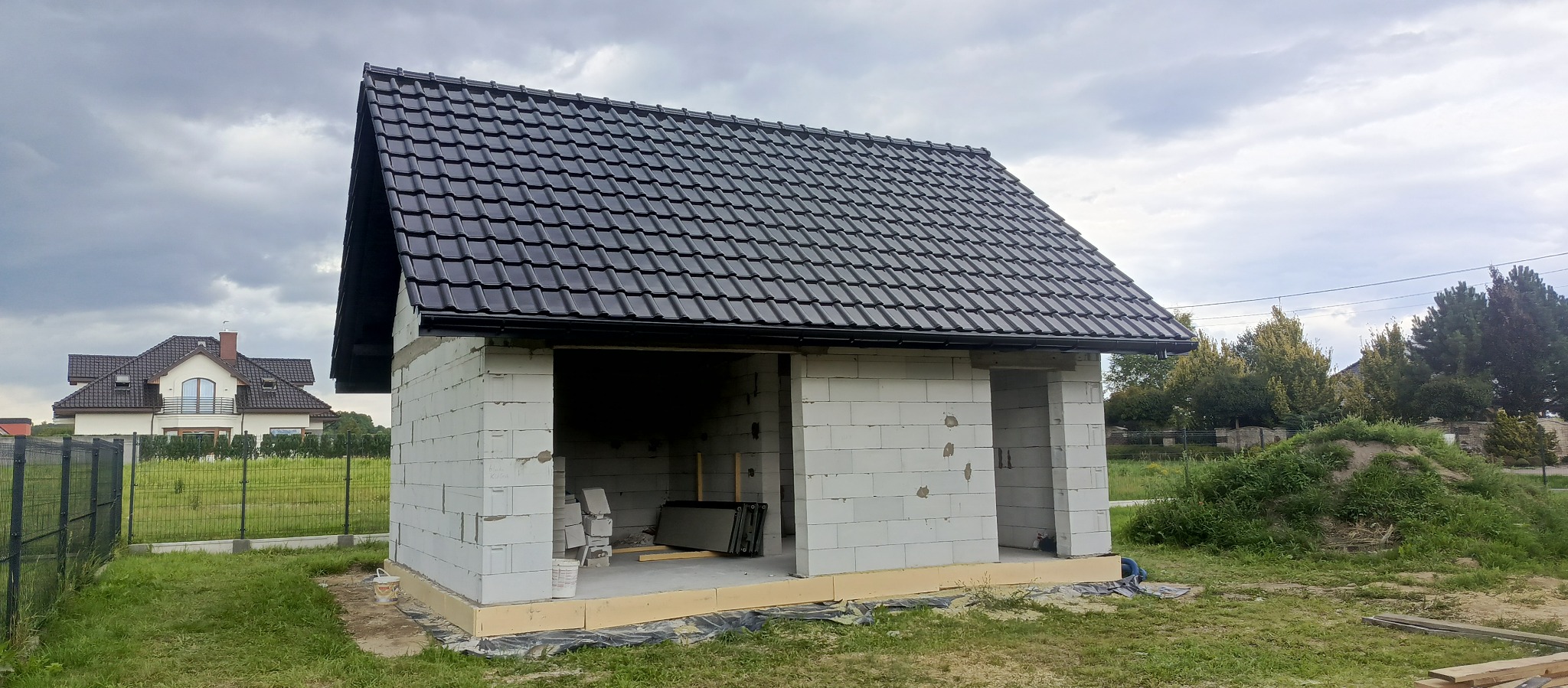 Niedokończony budynek gospodarczy z czarnym dachem z dachówki ceramicznej, ściany z białych bloczków, widoczne fundamenty, tło: dom jednorodzinny i zieleń.