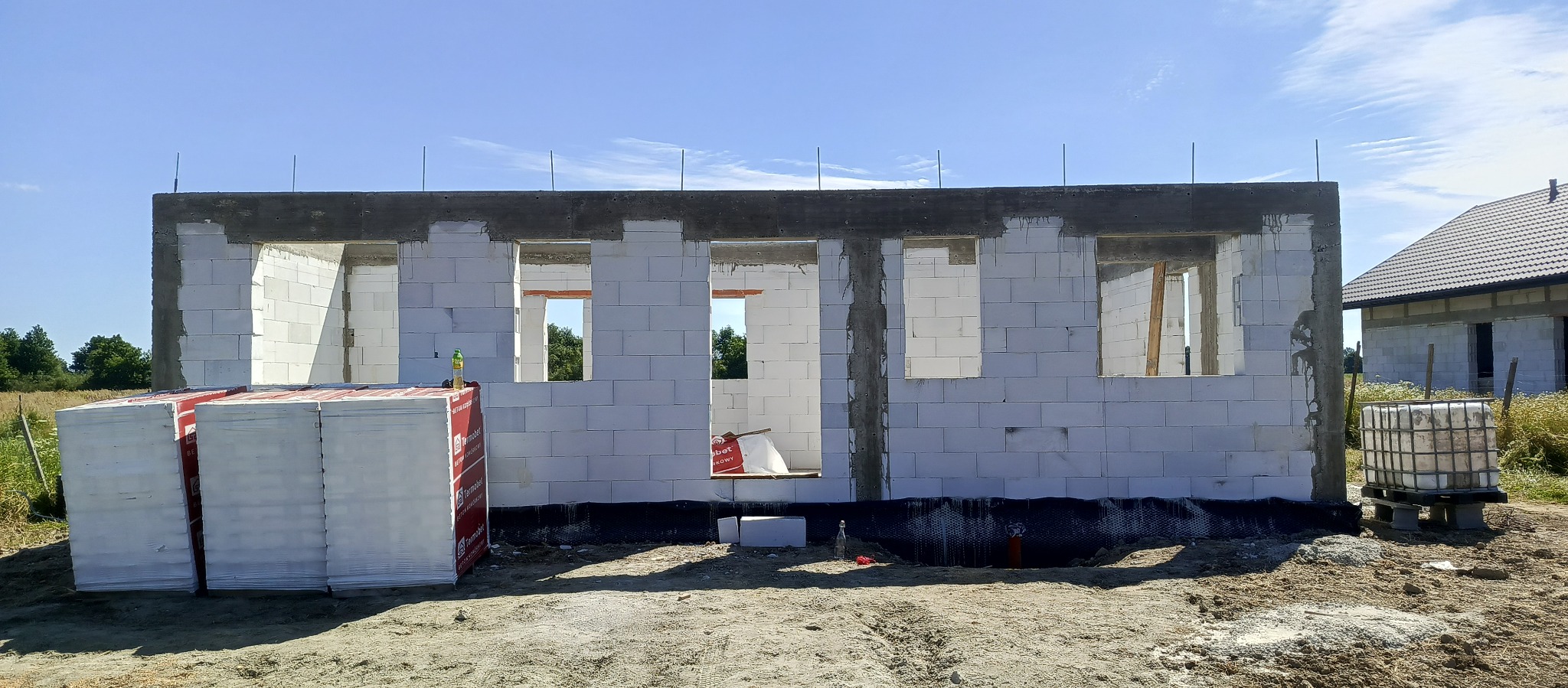 Wznoszenie ścian z białych bloczków betonu komórkowego. Widoczne otwory okienne i drzwiowe. Na pierwszym planie palety z materiałem budowlanym.