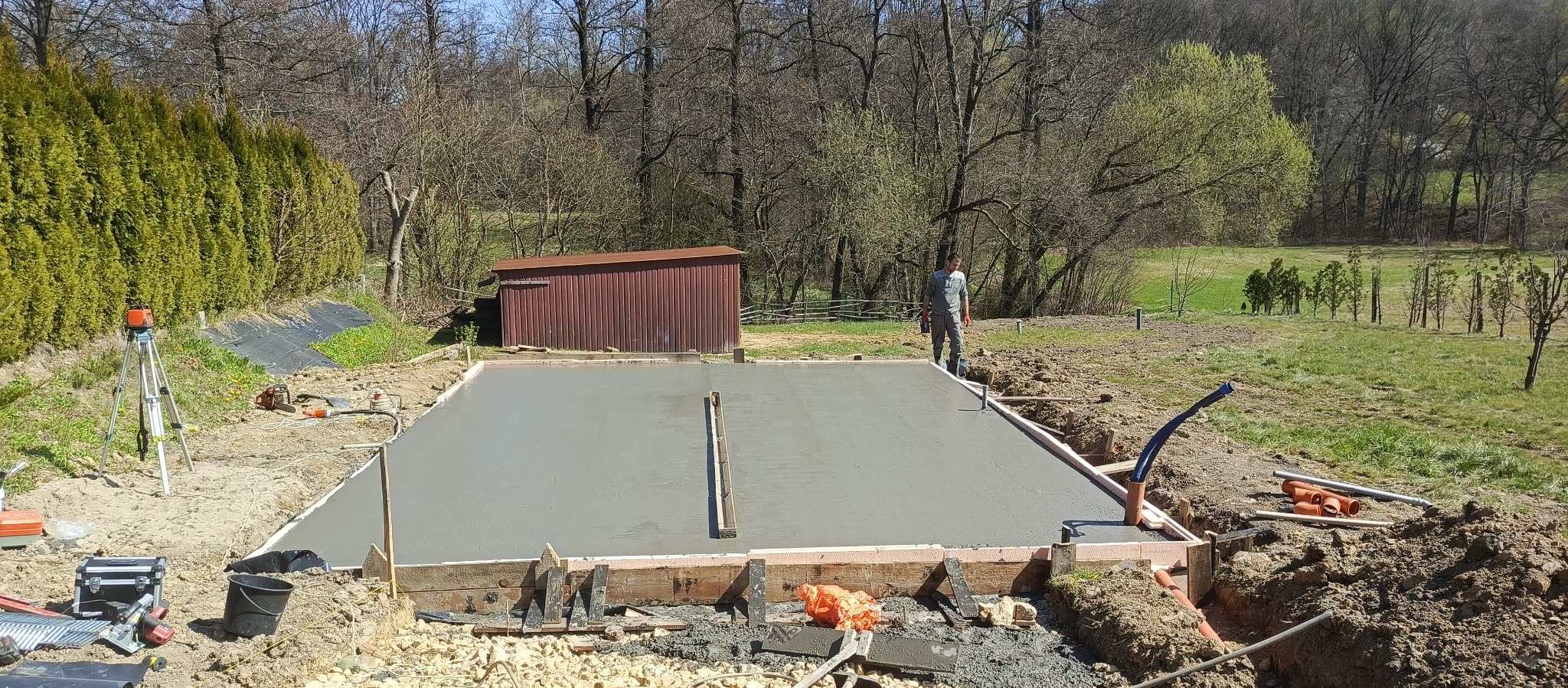 Wylany fundament pod budowę. Widoczne szalunki, narzędzia i pracownik na tle zieleni. Świeży beton, poziomowany łatą. Laser krzyżowy na statywie.