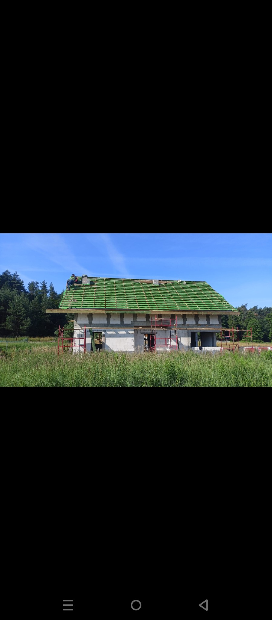 Niedokończony dom z zielonym dachem w trakcie budowy, widoczne rusztowania i ekipa na dachu, otoczony polami i lasem w słoneczny dzień.