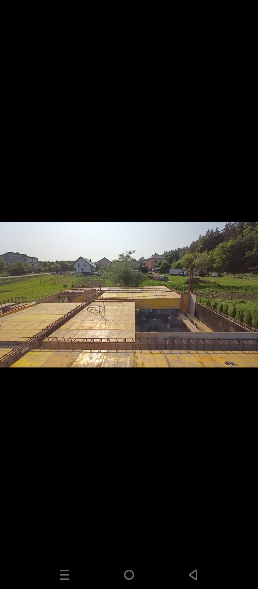 Wysoki kąt: szalunek fundamentów zbrojonych stalą na budowie domu, z widokiem na okoliczne domy i zieleń w tle, w słoneczny dzień.