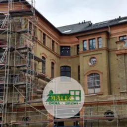 Budynek z żółtej cegły w trakcie montażu stolarki okiennej, widoczne rusztowania i zabezpieczone folią otwory okienne.