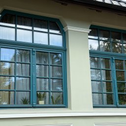 MALEC OKNA SPÓŁKA Z OGRANICZONĄ ODPOWIEDZIALNOŚCIĄ - Dwa okna drewniane w kolorze morskim z dekoracyjnymi szprosami i łukowym wykończeniem, widoczne rośliny doniczkowe wewnątrz, jasna elewacja budynku.