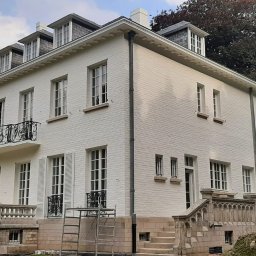 MALEC OKNA SPÓŁKA Z OGRANICZONĄ ODPOWIEDZIALNOŚCIĄ - Elewacja dwupiętrowego, białego domu z oknami i balkonem z kutą balustradą. Widoczne rusztowania i prace wykończeniowe. W tle drzewa i pochmurne niebo.