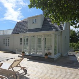 MALEC OKNA SPÓŁKA Z OGRANICZONĄ ODPOWIEDZIALNOŚCIĄ - Jasny, skandynawski dom z przeszkloną werandą i tarasem z leżakami. Widok z zewnątrz, słoneczny dzień, błękitne niebo. Charakterystyczna architektura.