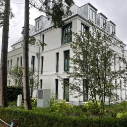 MALEC OKNA SPÓŁKA Z OGRANICZONĄ ODPOWIEDZIALNOŚCIĄ - Nowoczesny budynek mieszkalny z białymi ścianami i ciemnymi oknami, otoczony zielenią. Widoczne drzewa i krzewy w ogrodzie.